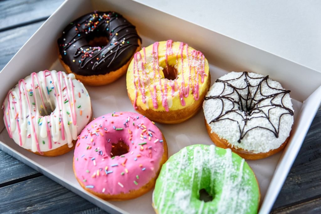 Caixa de delivery branca com seis donuts decorados com coberturas coloridas variadas e granulados sobre uma mesa de madeira. Ideias atrativas de sabores de donuts para vender.