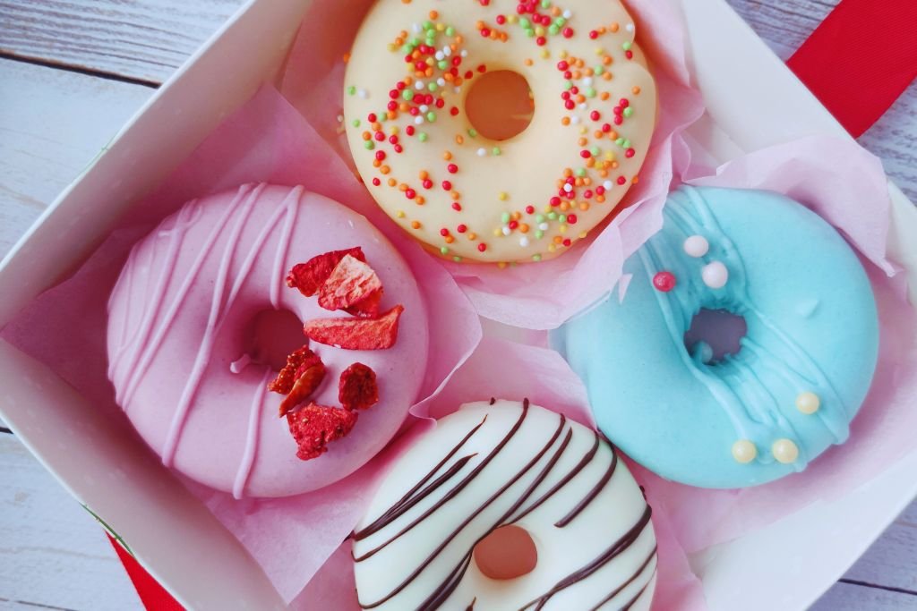 Caixa de presente branca com quatro donuts gourmet acomodados delicadamente em papel de seda rosa. Coberturas artesanais em tons pastéis (rosa com morangos desidratados, azul, amarelo com confeitos e branco com listras de chocolate). Ideia sofisticada de embalagem para vender donuts.