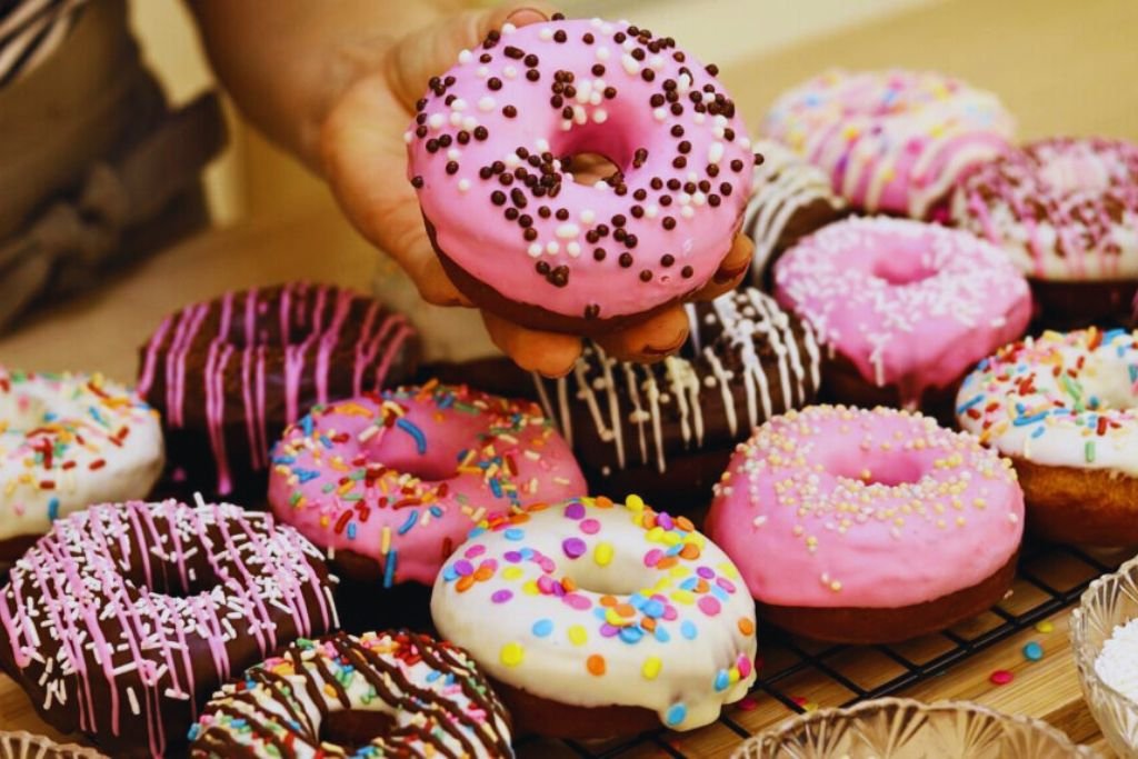 Mão segurando um donut fofinho com cobertura rosa e confeitos de chocolate. Ao fundo, vários donuts decorados em uma grade de resfriamento. Resultado final da receita de donuts para vender.